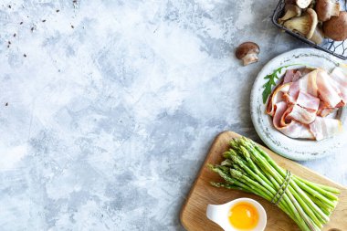 Gri beton arka planda yemek pişirmek için gerekli malzemeler. Bir demet taze kuşkonmaz, yumurta, pastırma, mantar. Üst Manzara. Uzayı kopyala Yatay