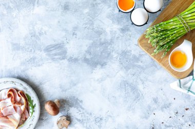 Gri beton arka planda yemek pişirmek için gerekli malzemeler. Bir demet taze kuşkonmaz, yumurta, pastırma, mantar. Üst Manzara. Uzayı kopyala Yatay