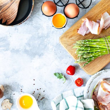 Gri beton arka planda yemek pişirmek için gerekli malzemeler. Bir demet taze kuşkonmaz, yumurta, pastırma, mantar, domates, roka. Üst Manzara. Uzayı kopyala Kare