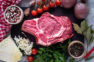 Çiğ sulu biftek, siyah tahtada ızgara yapmaya hazır. Kemikte biftek, ahşap kesim tahtasında kiraz domatesleri, acı biber, kırmızı fasulye ve bitkilerle birlikte dana eti. Borç için malzemeler. en üstteki görünüm.