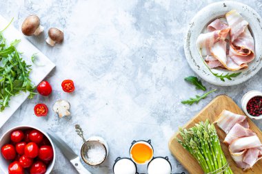 Gri beton arka planda yemek pişirmek için gerekli malzemeler. Bir demet taze kuşkonmaz, yumurta, pastırma, mantar, domates, roka. Üst Manzara. Uzayı kopyala Yatay