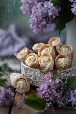 Baharda metal bir kutuda vanilya kremalı poğaça boynuzları ve ahşap bir masada bir buket leylak..