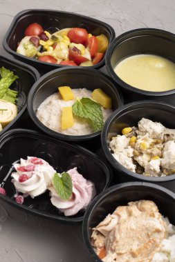 Plastik kaplarda lezzetli öğle yemeği, bıldırcın yumurtalı krema çorbası, arpa lapası ve gri masada vegan salatası.