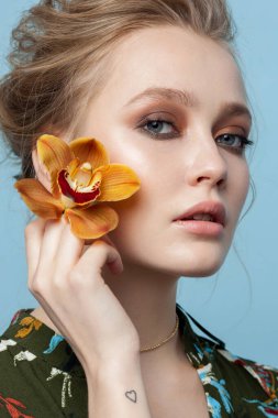 Yüzüne yakın orkidesi olan genç bir manken, cilt bakımı konsepti, güzellik çekimi, yakın plan çekici kadın portresi, parlak makyaj, sarı saç.