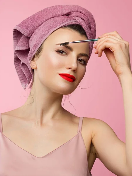 Close-up de uma mulher bonita com maquiagem natural e pele limpa com uma toalha na cabeça. Menina penteando sobrancelhas grossas e olhando para a câmera. Cosmetologia e spa. Hora da manhã e maquiagem — Fotografia de Stock