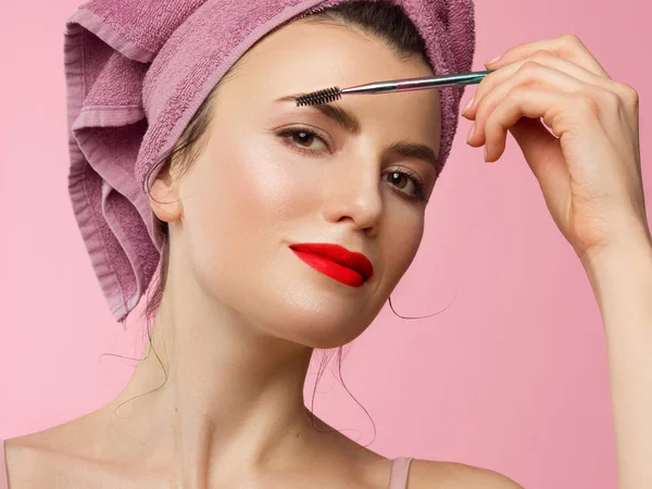 Primer plano de una hermosa mujer con maquillaje natural y piel limpia con una toalla en la cabeza. Chica peinando cejas gruesas y mirando a la cámara. Cosmetología y spa. Mañana y maquillar. LI rojo —  Fotos de Stock