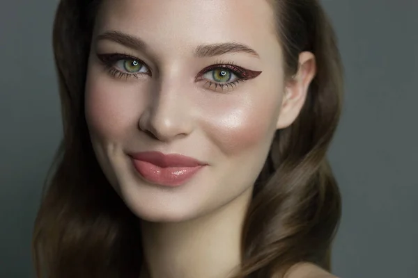 Vacker brunett modell kvinna ansikte med gröna ögon och perfekt make-up gör leende på kameran. Porträtt av skönhet med naturliga läppar och eyeliner, tjocka ögonbryn. Kvinnligt ansikte med klar hud närbild. — Stockfoto