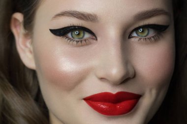 Yeşil gözlü, güzel esmer model kadın yüzü ve mükemmel makyajı kamerada gülümsemesini sağlıyor. Güzel bir portre, kırmızı dudaklar ve göz kalemi, kalın kaş. Açık tenli bir kadın yüzü. Yakın plan..