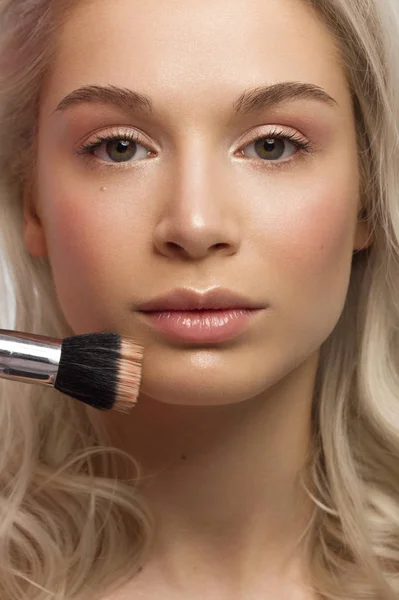 Schönheit Nahaufnahme einer blonden Frau, die Puder aufträgt, um ihre Gesichtshaut zu reinigen. blickt mit einem sexy Blick in die Kamera. Weiche Hautpflege, natürliches Make-up. rosa Lipgloss, glatte und dicke Augenbrauen. lockiges Haar — Stockfoto