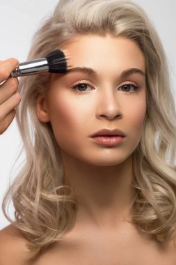 Sarışın bir kadının cildini temizlemek için pudra sürerken güzel bir fotoğrafı. Seksi bir bakışla kameraya bakıyor. Yumuşak cilt bakımı, doğal makyaj. Pembe dudak parlatıcısı, pürüzsüz ve kalın kaşlar. kıvırcık saçlı