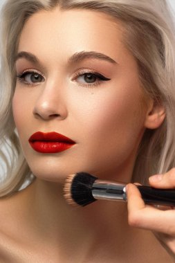 Sarışın bir kadının cildini temizlemek için pudra sürerken güzel bir fotoğrafı. Seksi bir bakışla kameraya bakıyor. Yumuşak cilt bakımı, doğal makyaj. Pembe dudak parlatıcısı, pürüzsüz ve kalın kaşlar. kıvırcık saçlı