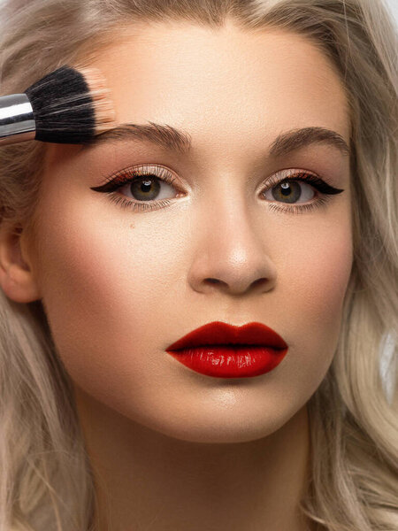 Красота крупным планом блондинка применяет порошок, чтобы очистить кожу лица. Смотрит в камеру с сексуальным взглядом. Мягкий уход за кожей, натуральный макияж. Блеск для губ розовый, гладкие и толстые брови. вьющиеся волосы
