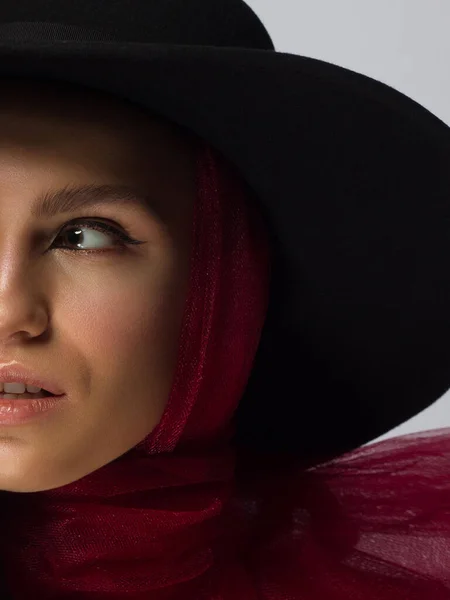 Half face beauty of a woman with a hat, black eyeliner and a red scarf. Clean and shine skin, natural lip gloss, thick eyebrows and long eyelashes. Vintage photography, cosmetology and makeup.