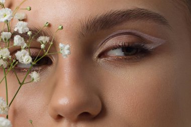 Çıplak makyajlı bir kadının gözlerine yakın çekim. Modaya uygun göz farı, son derece uzun kirpikleri ve kalın pürüzsüz kaşları var. Güzel bir görünüş. Temiz ten ve beyaz çiçekler.