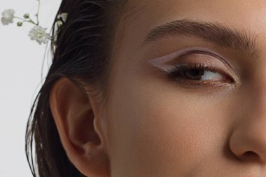 Çıplak makyajlı bir kadının gözlerine yakın çekim. Modaya uygun göz farı, son derece uzun kirpikleri ve kalın pürüzsüz kaşları var. Güzel bir görünüş. Temiz ten ve beyaz çiçekler.