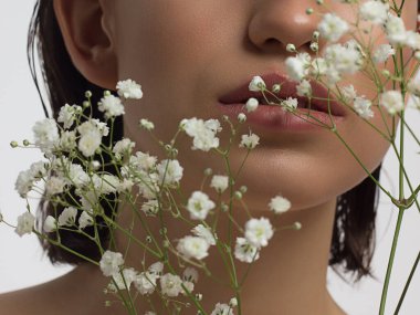 Güzel ciltli ve çıplak makyajlı kadın dudaklarının makro fotoğrafı. Dolgun dudaklarda şeffaf bir parlatıcı ve beyaz çiçekler. Moda fotoğrafçılığı. Kozmetik ve kaplıca. Baharın tazeliği