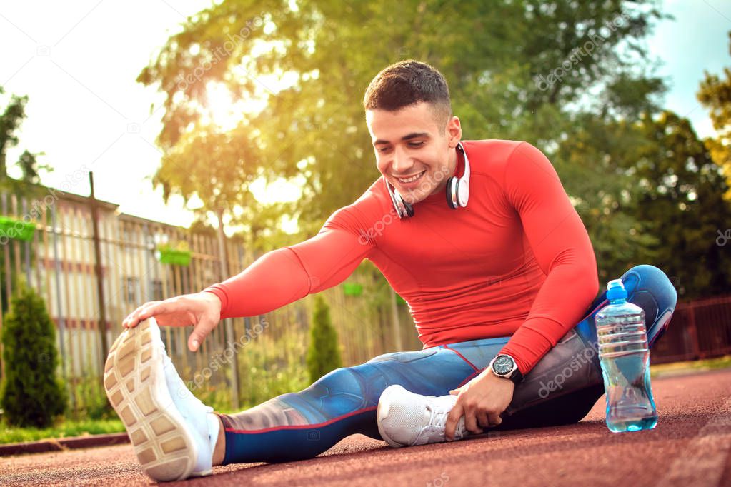 Handsome athlete is sitting on the road. He is stretching one leg forward while another leg is bent. He stretches himself with seriousness.