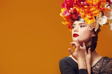 Sanat güzellik portresi. Turuncu arka planda parlak turuncu şapkalı ve kafasında klasik orkideler olan muhteşem genç bir kadın. Bahar ve yaz güzelliği. Parfüm. Moda..