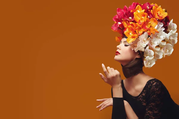 Art beauty portrait. Gorgeous young woman in a hat of bright orange and vinous orchids on her head on an orange background. Spring and summer beauty. Perfume. Fashion.