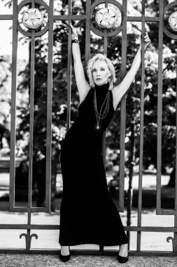 Çitin yanındaki yaz parkında yaşlı, şık bir kadının tam boy siyah beyaz portresi. Güzellik, moda.