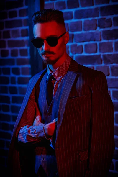 Portrait of a handsome young man in elegant suit and black sunglasses standing in red light by a brick wall. Men\'s fashion. Luxurious lifestyle.