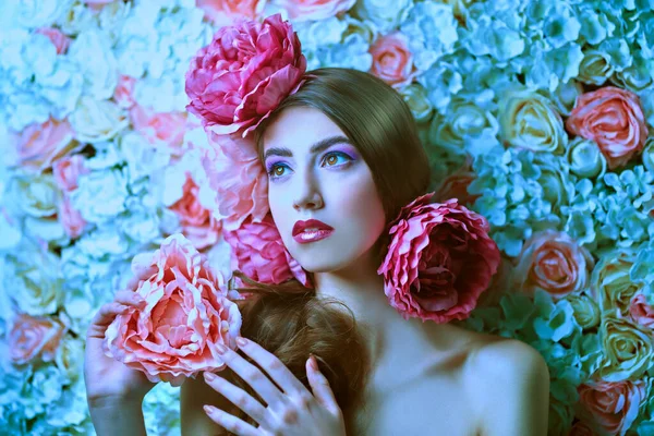Beautiful young woman with gentle purple lilac make-up and peony flowers in her hair posing on a background of roses. Inspiration of spring and summer. Perfume, cosmetics concept.