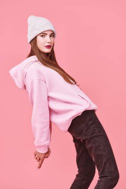 Gençlik modası. Pembe kazaklı, şapkalı, stüdyoda pembe arka planda poz veren modern bir genç kız. Kozmetik ürünleri ve makyaj.