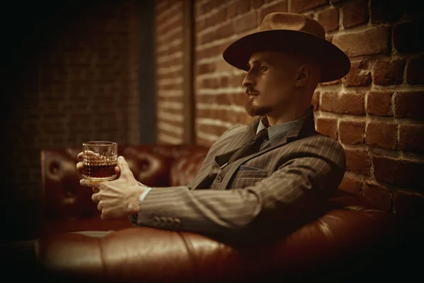 Handsome confident man sitting relaxed in a rich setting on a leather sofa with a glass of whiskey. Luxurious lifestyle. Retro style. Mafia and criminal world.