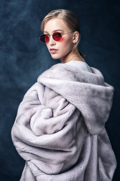 A portrait of a young fashionable woman in a fur coat with hood and sunglasses. Beauty, optics, fashion.
