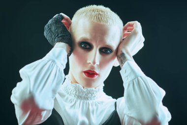 Kadın makyajlı ve kadın kıyafetleriyle stüdyoda ifade ile poz veren genç bir adamın tam boy portresi..