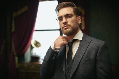 Yakışıklı beyefendi tüvit takım elbise ve gözlüklerle poz veriyor ve gömleğinin yakasını eski moda bir iç mekâna göre ayarlıyor. Lüks bir yaşam tarzı. Retro biçimi. 