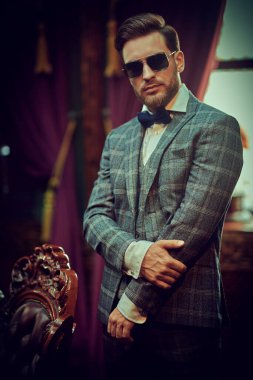Handsome gentleman poses in a checked suit and sunglasses in a vintage interior. Luxurious lifestyle. Retro style.  clipart
