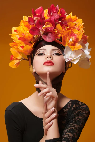 Art beauty portrait. Gorgeous young woman in a hat of bright orange and vinous orchids on her head on an orange background. Spring and summer beauty. Perfume. Fashion.
