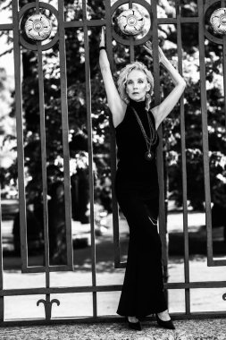 Çitin yanındaki yaz parkında yaşlı, şık bir kadının tam boy siyah beyaz portresi. Güzellik, moda.