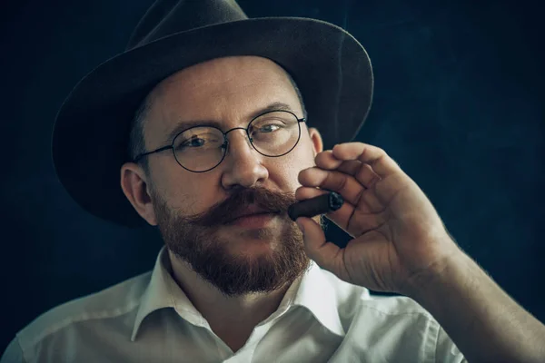 A close up portrait of a thoughtful man with a cigar posing in the studio over the black background. Men's beauty, fashion, style.