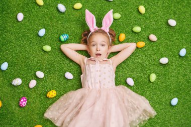 Renkli Paskalya yumurtaları arasında bir çim üzerinde yalan tavşan kulaklı küçük kız. Paskalya tatili.