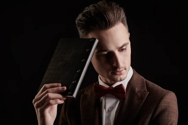 Portrait of a handsome man in elegant classic suit and bow tie with a leather diary on a black background. Business style. Men\'s fashion.
