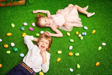 Neşeli küçük bir çocuk ve tavşan kulaklı bir kız renkli yumurtalarla çimlerin üzerinde yatıyorlar. Paskalya tatili.