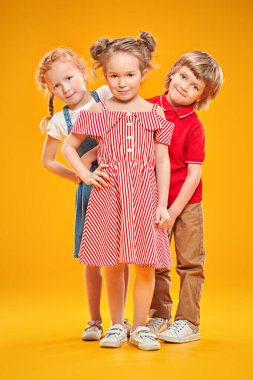 İki kız ve parlak yaz elbiseli bir oğlan stüdyoda sarı arka planda poz veriyorlar. Yaz çocuk modası. Tam boy portre.