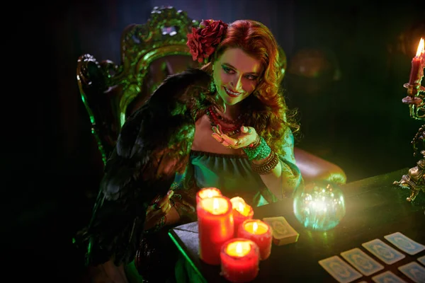 A beautiful red-haired fortune teller is talking to a black raven. The atmosphere of magic and sorcery, magic crystal ball and tarot cards.