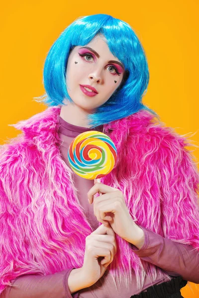 Attractive party girl with bright pink makeup and blue wig wears pink fur coat and holds big lollipop on a yellow background. Make-up and cosmetics, hairstyle. Fashion girl.