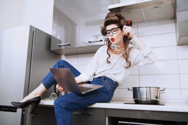 Saçında bigudiler olan güzel bir kız elinde bir kepçe ve dizüstü bilgisayarla günlük kıyafetleriyle poz veriyor. Moda ev çekimi. Pin-up tarzı.