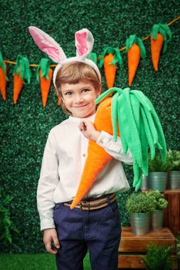 Tavşan kulaklı mutlu küçük çocuk bahar bahçesinde büyük bir havuç ve Paskalya süslemeleri duruyor. Paskalya tatili.
