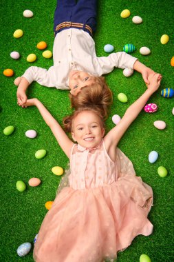 Şirin, gülümseyen bir oğlan ve tavşan kulaklı bir kız renkli yumurtalarla yeşil bir çimenlikte uzanıyorlar. Paskalya tatili.