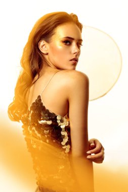 Altın makyaj malzemesi. Altın makyajlı ve arka planda parlak altın rengi elbisesi olan güzel bir genç kadın. Parti biçimi.