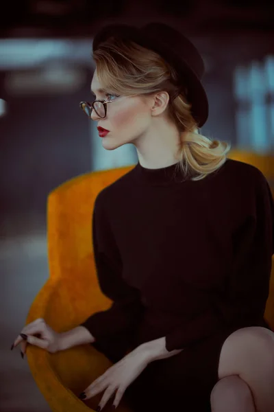 Beautiful fashionable lady in elegant black dress, a hat and glasses sitting in modern yellow armchair. Beauty, fashion. Optics style.