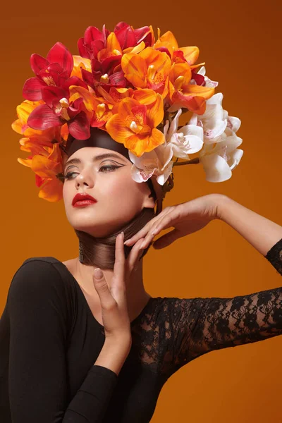 Art beauty portrait. Gorgeous young woman in a hat of bright orange and vinous orchids on her head on an  orange brown background. Spring and summer beauty. Perfume. Fashion.