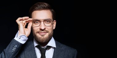 Zarif takım elbiseli ve kameraya gülümseyen gözlüklü yakışıklı bir adam. Siyah arka planda bir iş adamı portresi. Erkek güzelliği, moda. Erkekler için optik.