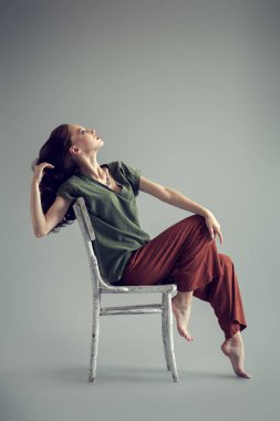 Stüdyonun gri arka planında sandalyede poz veren güzel bir genç kadının tam boy portresi. Sıradan moda, fitness, yoga.