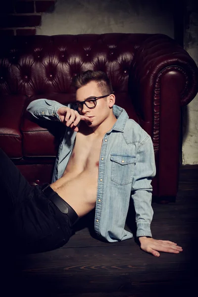 Attractive young man in a denim shirt with a naked torso sits on the floor next to a leather sofa. Men\'s beauty, fashion.